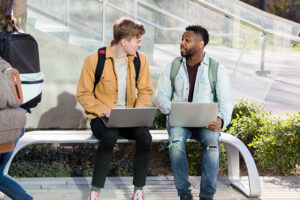 two male college students sitting on a bench outside talking to one another while holding laptops