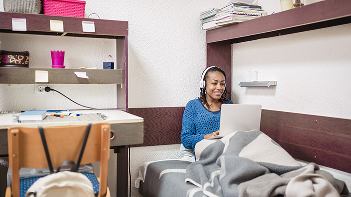 Young African female student, relaxing in a University campus room. She listening music and checking social media using laptop and mobile phone